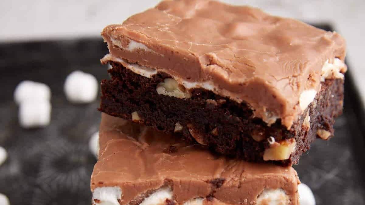 A close-up of two stacked chocolate brownies with a layer of chocolate frosting and visible marshmallows and nuts, placed on a dark surface with a few mini marshmallows scattered nearby.