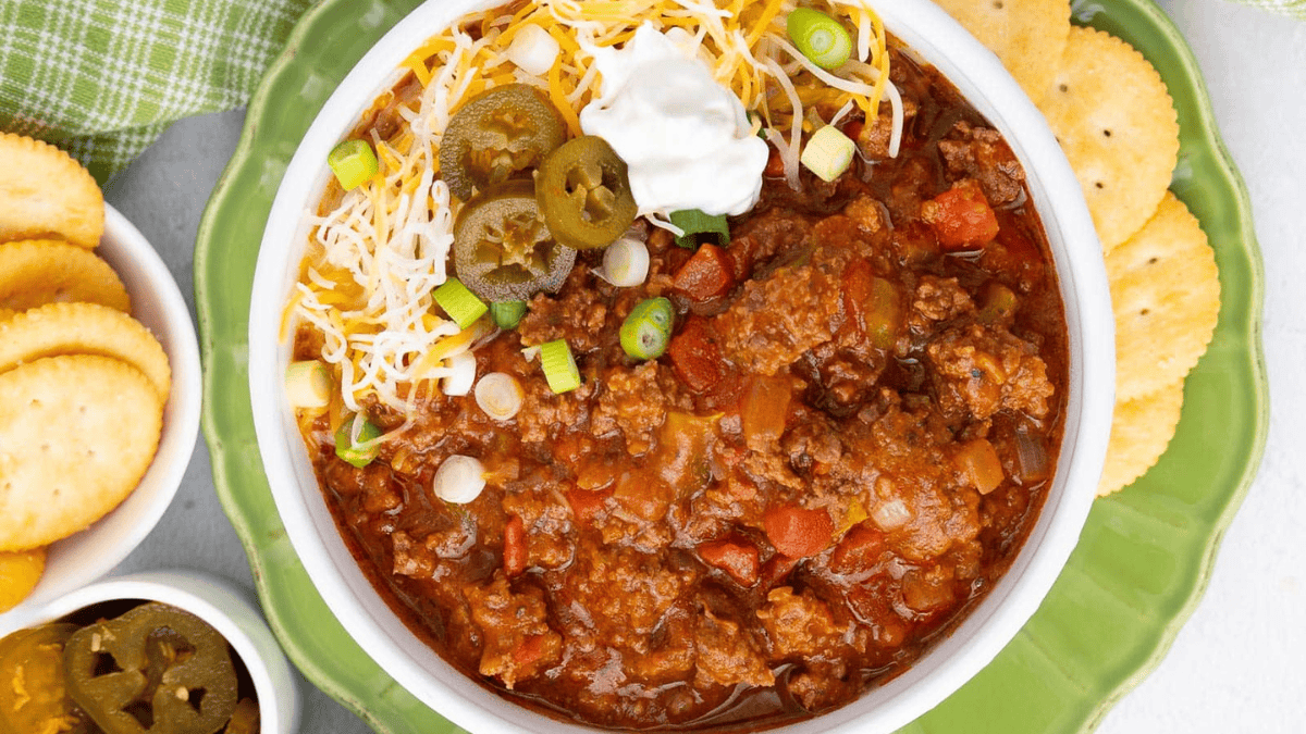 A bowl of chili topped with shredded cheese, green onions, jalapeños, and sour cream is served with Ritz crackers on a green plate. Don’t waste your slow cooker’s potential—let it create this flavorful meal while you relax.