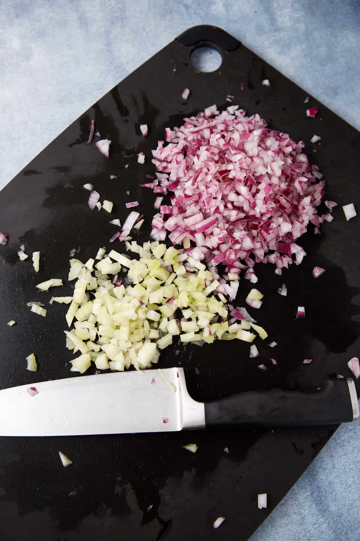 A black cutting board with finely chopped red onion and celery, a chef's knife lying beside the piles of chopped vegetables.