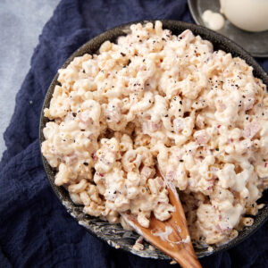 A bowl of creamy macaroni salad with visible bits of onion, topped with cracked black pepper, sits on a dark cloth. A wooden spoon is partially submerged in the salad.