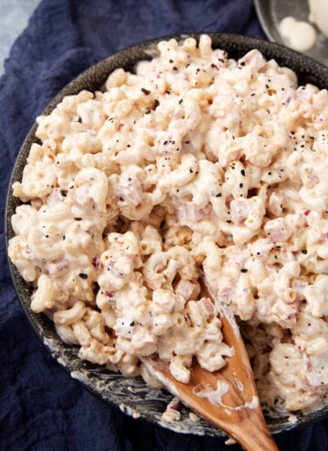 A bowl of creamy macaroni salad with visible bits of onion, topped with cracked black pepper, sits on a dark cloth. A wooden spoon is partially submerged in the salad.