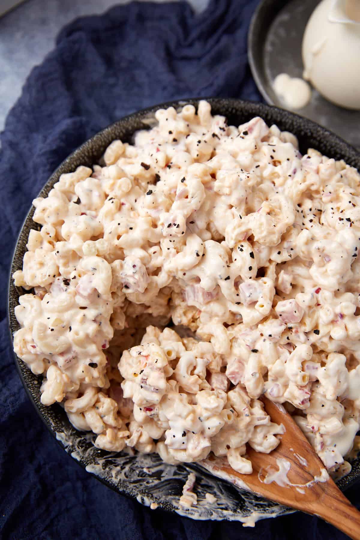 A close-up of creamy macaroni salad with diced ham and black pepper, served in a dark bowl with a wooden spoon, set on a dark blue cloth.