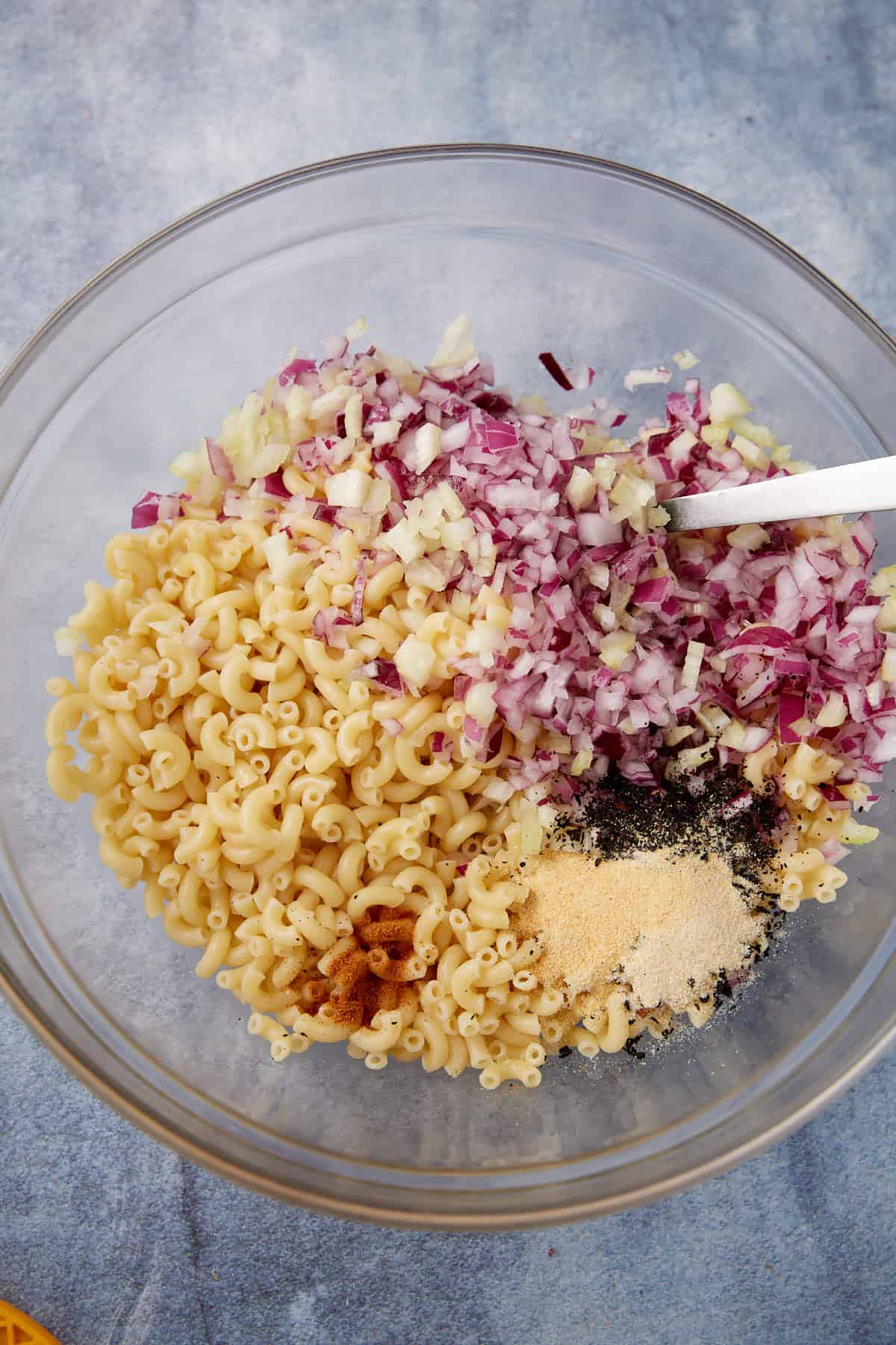 A glass bowl containing cooked elbow macaroni, finely chopped red onions, minced garlic, and various powdered spices, with a white spoon resting on the side.