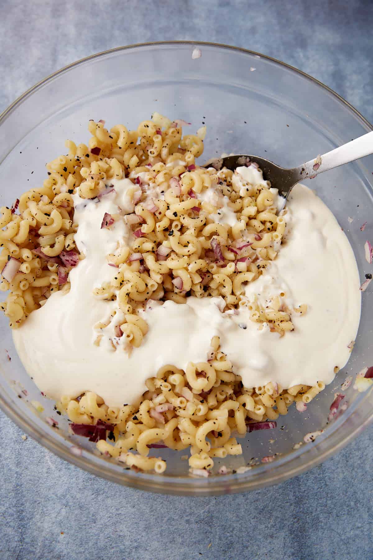 A glass bowl filled with cooked elbow macaroni, chopped red onions, black pepper, and a creamy white sauce being mixed with a large spoon.
