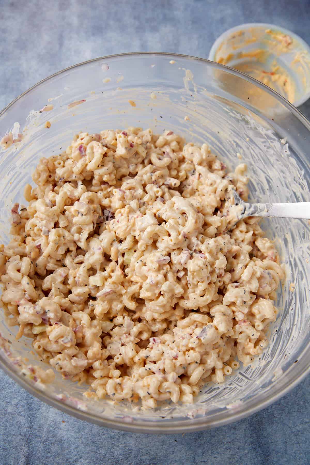 A glass bowl filled with creamy macaroni salad, mixed with small bits of ingredients, and a metal spoon. In the background, there’s a small bowl with leftover sauce. The scene is on a blue surface.