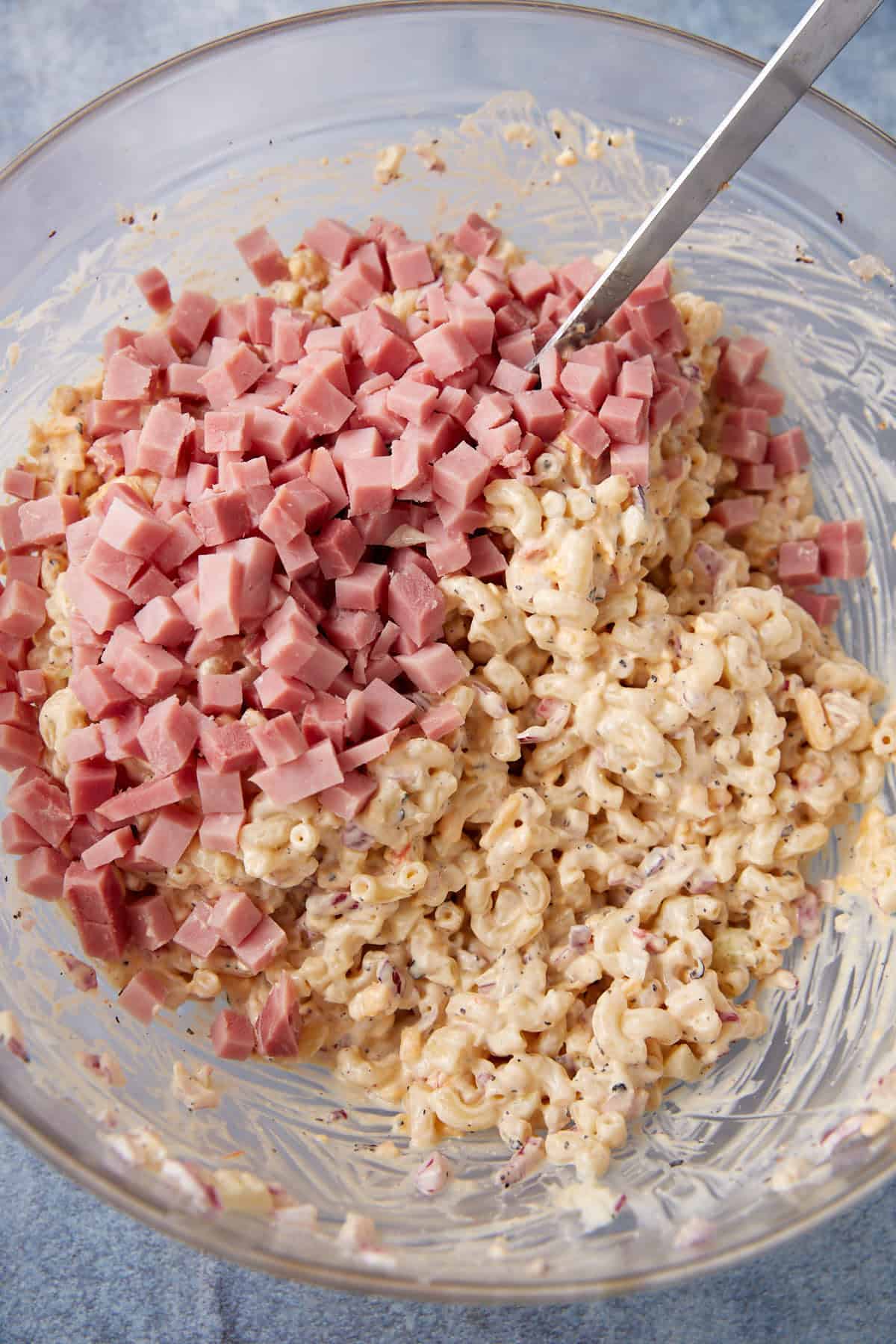 A glass bowl filled with creamy macaroni salad, topped with a pile of diced ham, and a metal spoon resting inside the mixture.