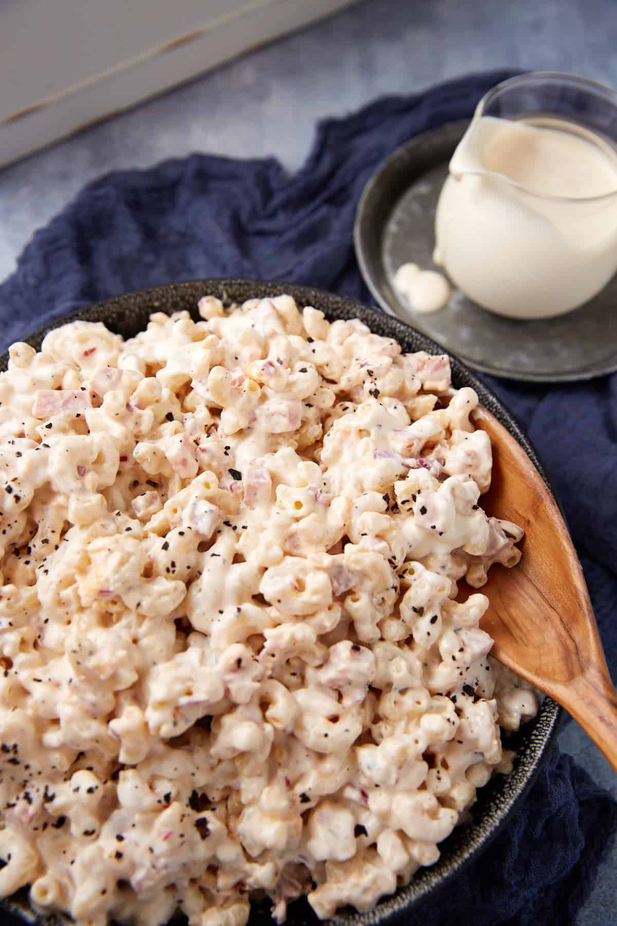 A bowl of creamy macaroni salad with a wooden spoon, topped with black pepper, sits on a dark cloth. A small pitcher of extra dressing is placed on a plate beside the bowl.