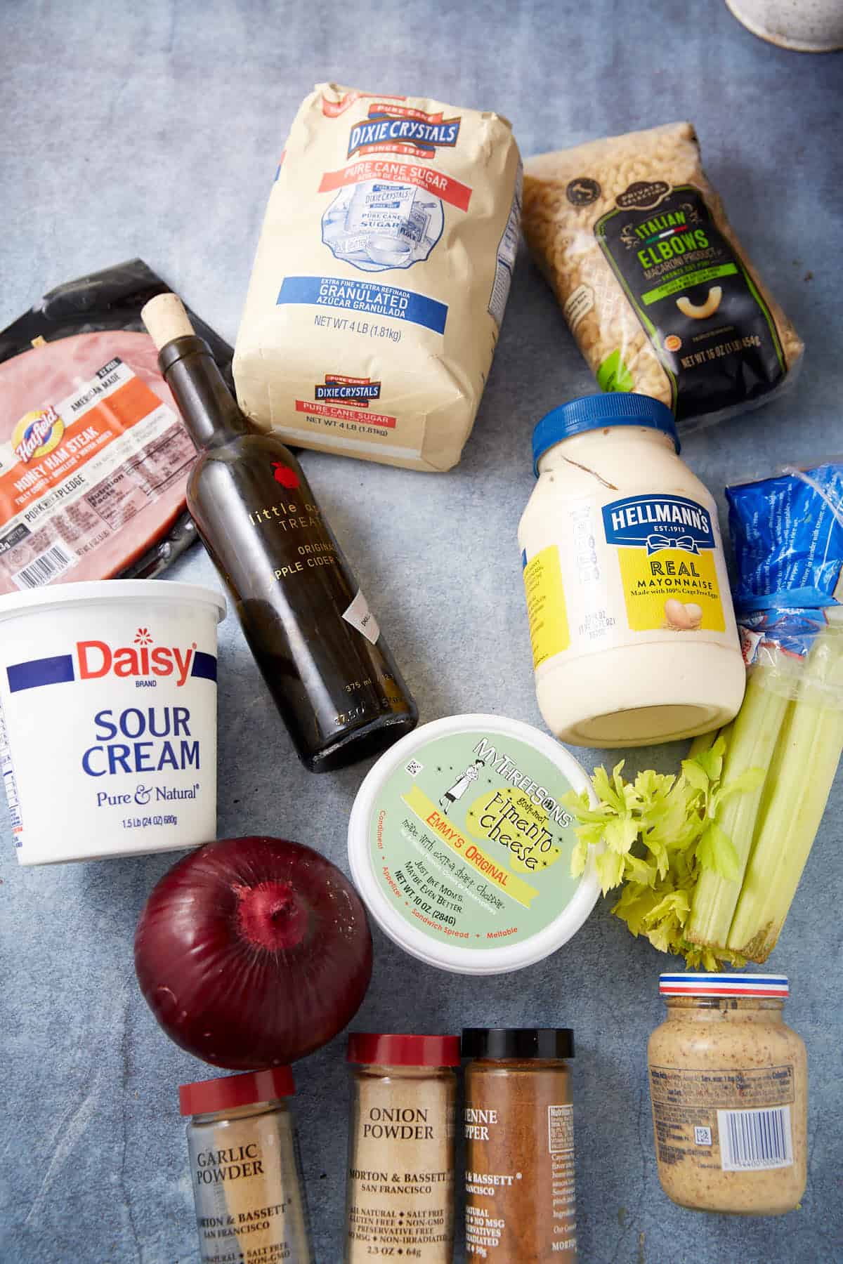 Various ingredients on a blue surface, including a bag of sugar, a jar of mayonnaise, sour cream, a red onion, celery, deli ham, canned chickpeas, balsamic vinegar, dry spices, Dijon mustard, and a block of cheese.