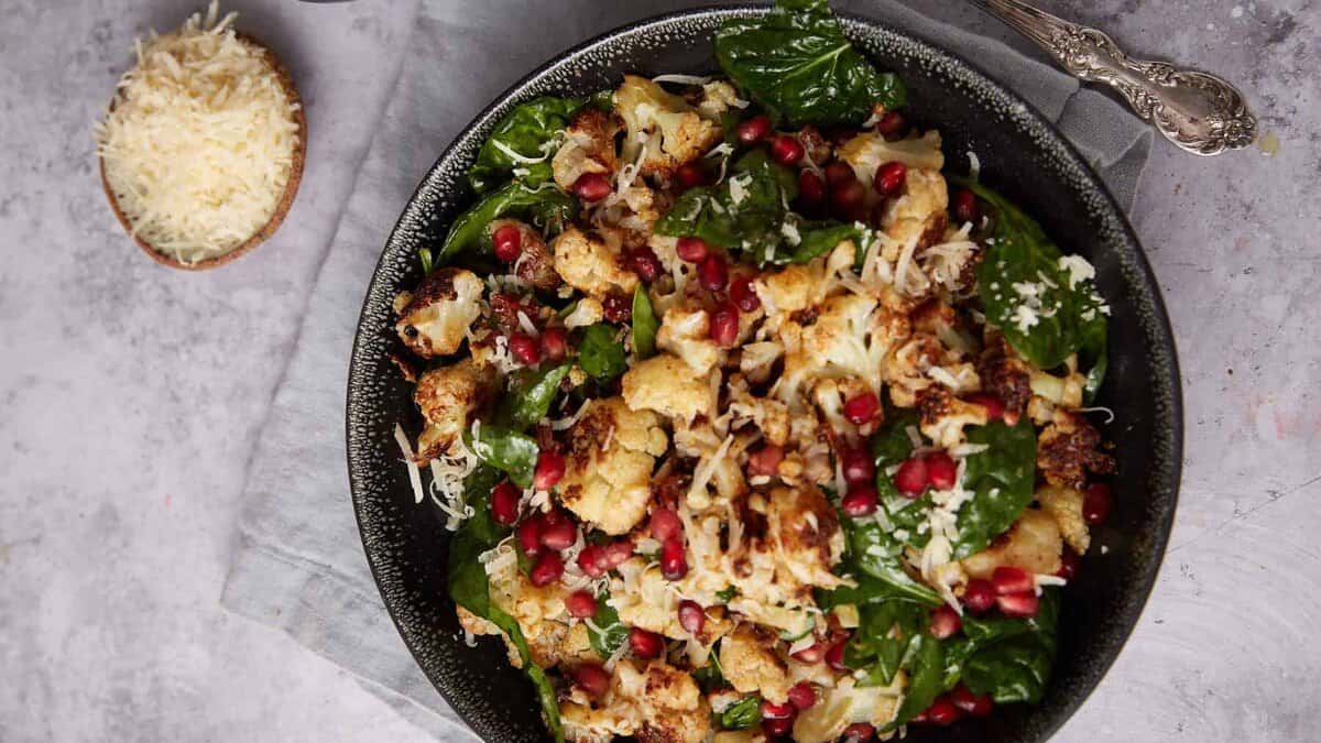 A bowl of salad with roasted cauliflower, fresh spinach leaves, shredded cheese, and pomegranate seeds, placed on a gray napkin next to small bowls of grated cheese and pomegranate seeds, with a spoon beside it.