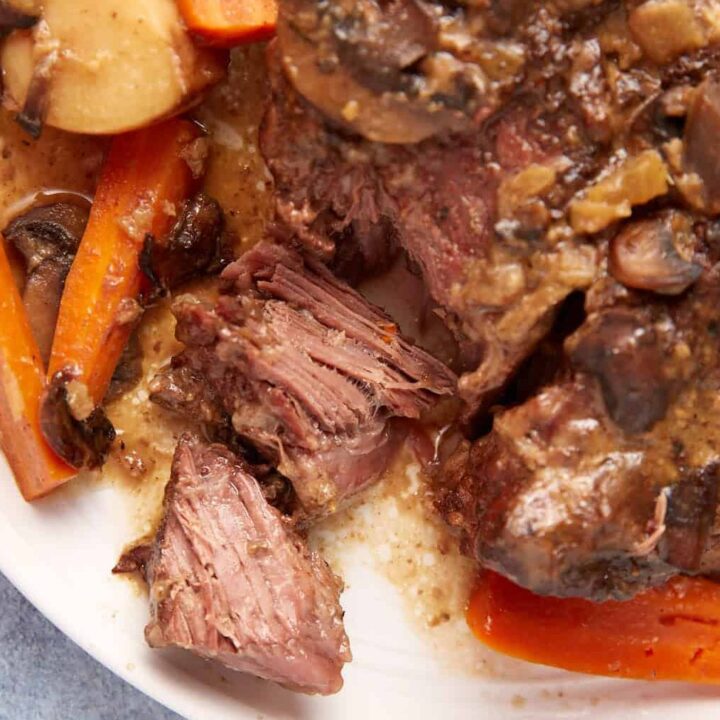 A close-up of a plate of slow cooker pot roast with gravy, featuring tender, shredded beef, carrots, mushrooms, and potatoes in a savory sauce. The dish is placed on a white plate against a light blue tablecloth background.