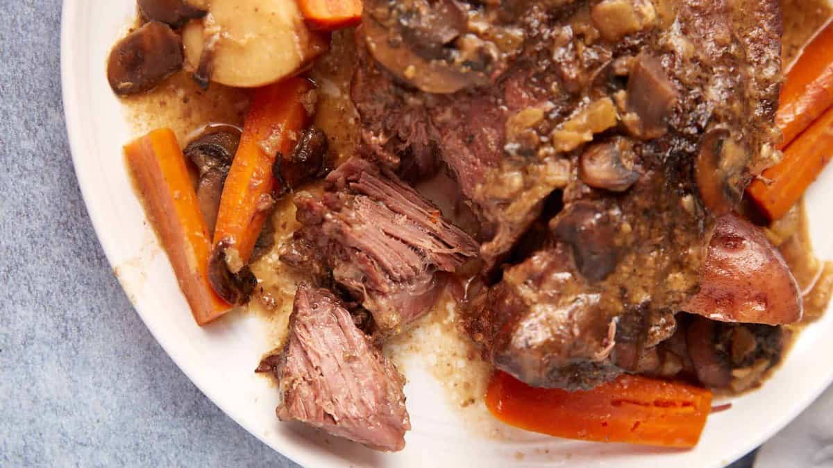 A close-up of a plate of slow cooker pot roast with gravy, featuring tender, shredded beef, carrots, mushrooms, and potatoes in a savory sauce. The dish is placed on a white plate against a light blue tablecloth background.