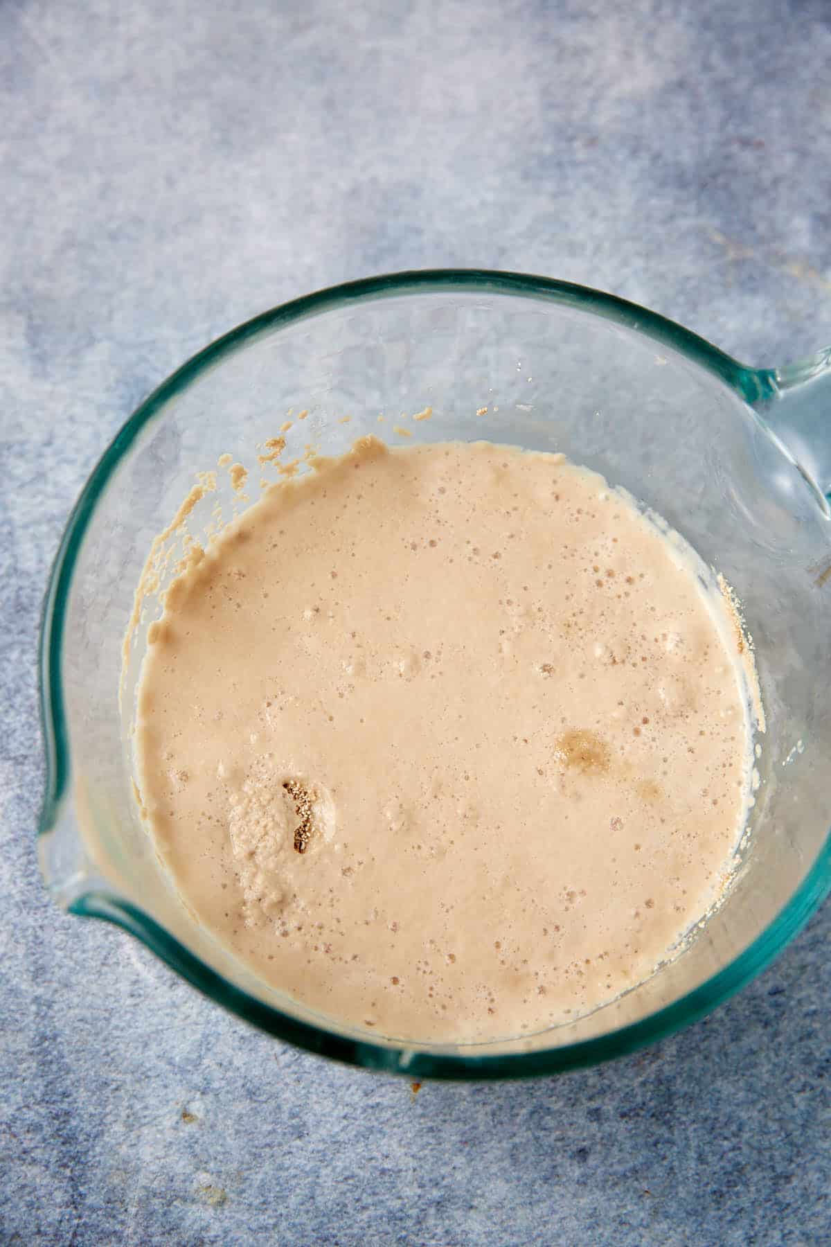 A glass measuring cup filled with bubbly, activated yeast mixture for soft pretzels sits on a light blue surface.