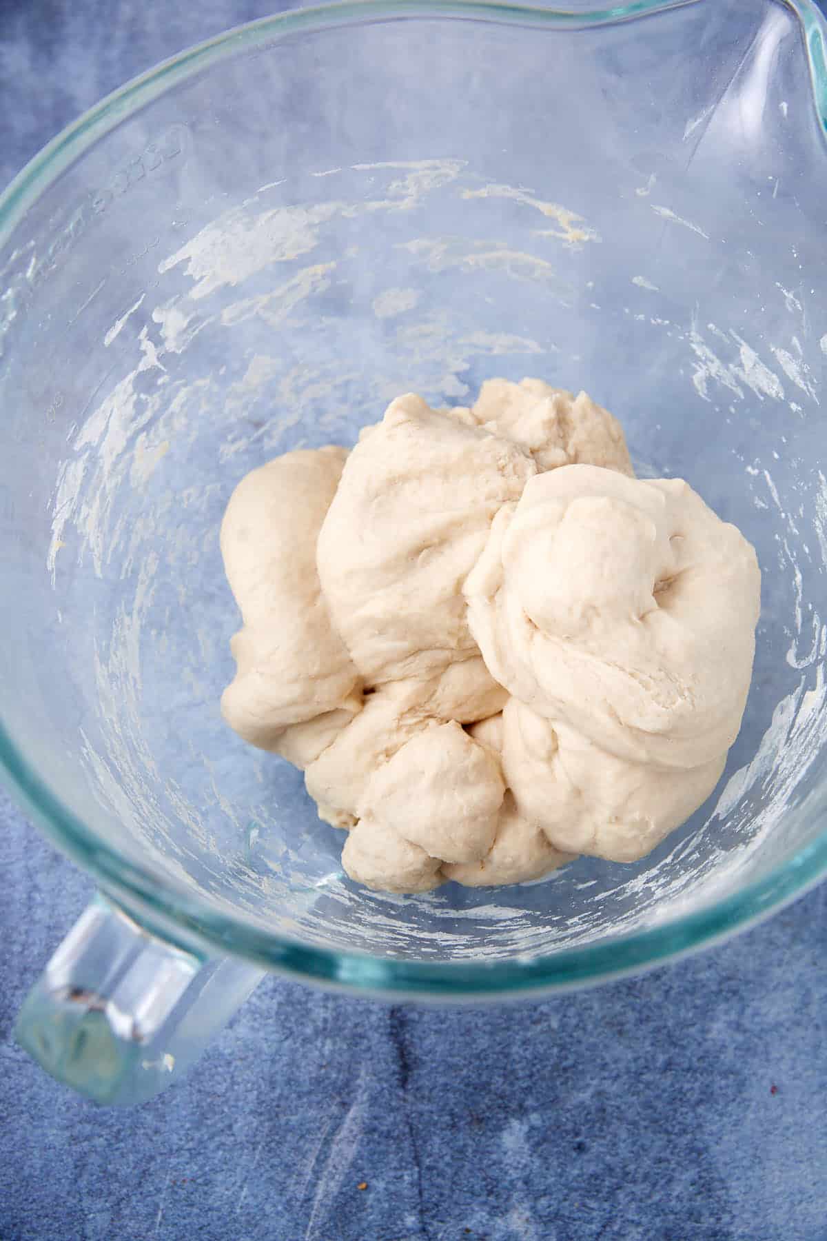 A ball of raw soft pretzel dough sits in a clear glass mixing bowl on a blue countertop.