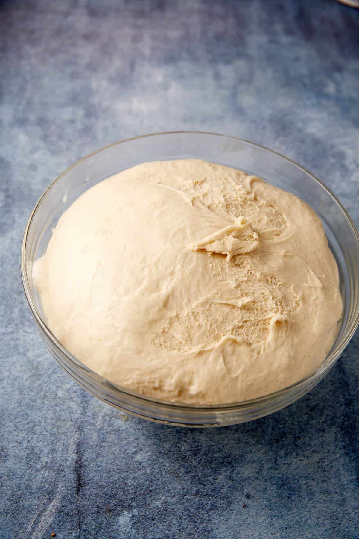 A glass bowl filled with risen bread dough for soft pretzels sits on a blue textured surface. The dough is smooth and puffy, with a few cracks on top.