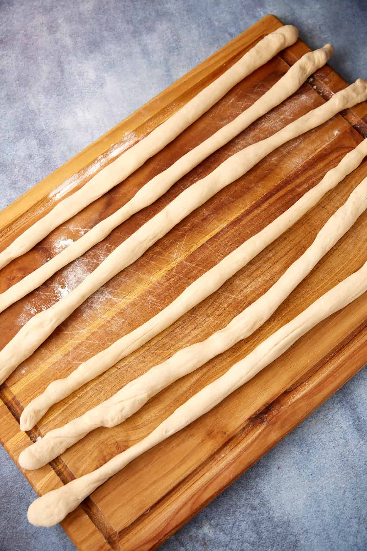 Six long, thin ropes of dough—perfect for shaping into soft pretzels—are laid out in parallel lines on a lightly floured wooden cutting board, set atop a blue countertop.