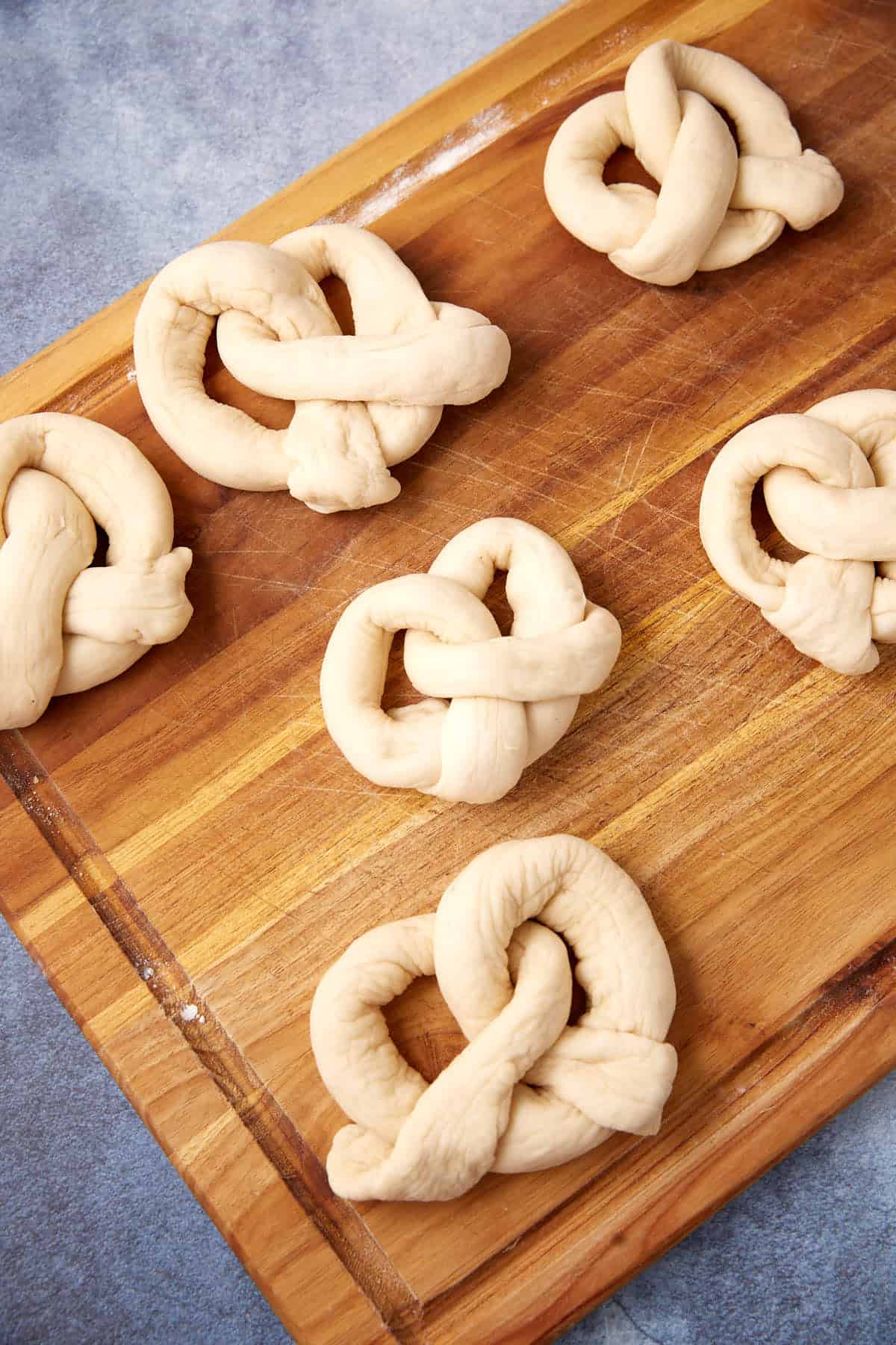 Six soft pretzels made from twisted dough sit unbaked on a wooden board, ready for the oven. The background features a textured blue surface.