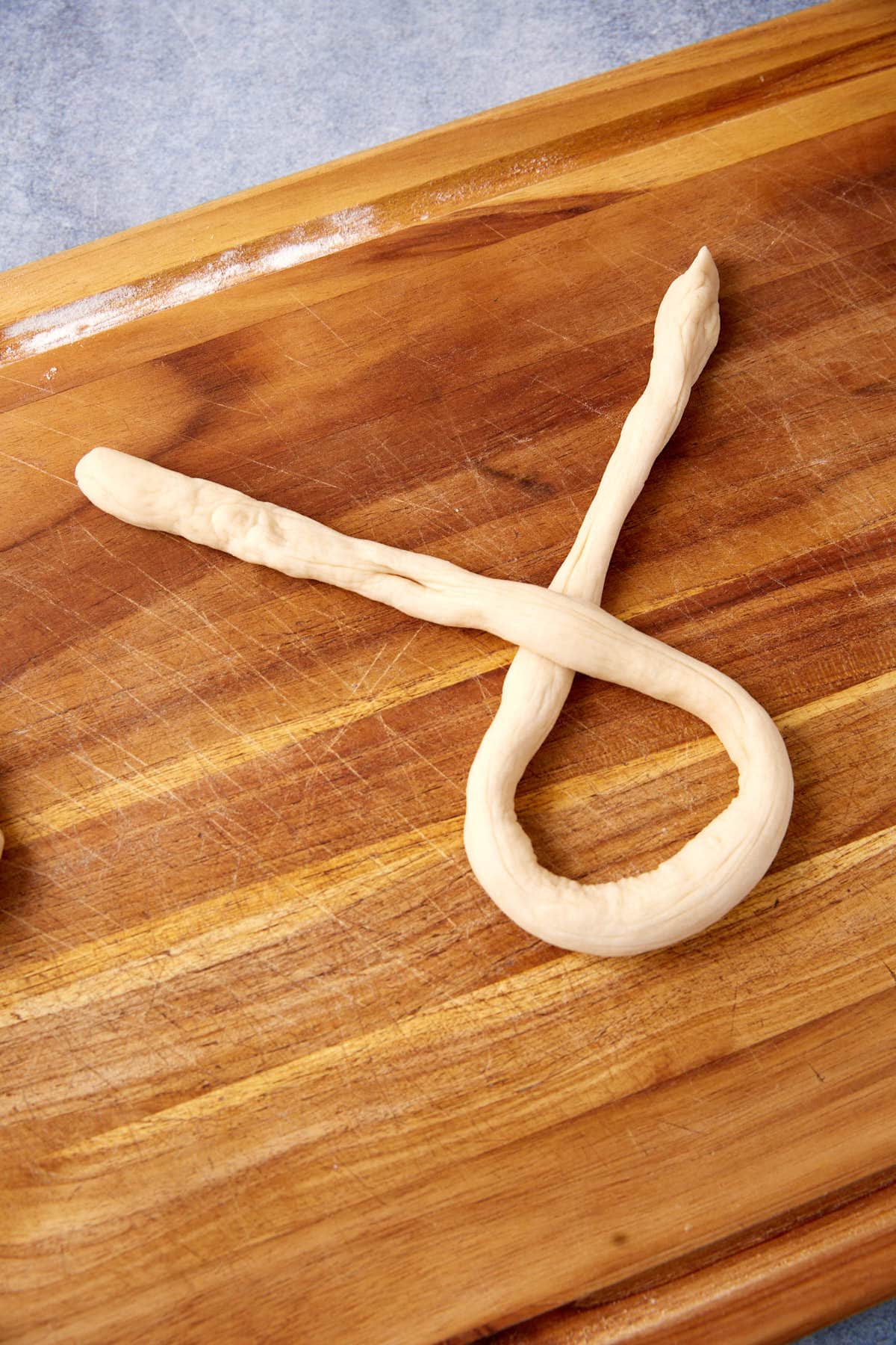 A strand of dough is twisted into a loop, one end crossing over the other on a wooden cutting board, preparing to shape classic soft pretzels.