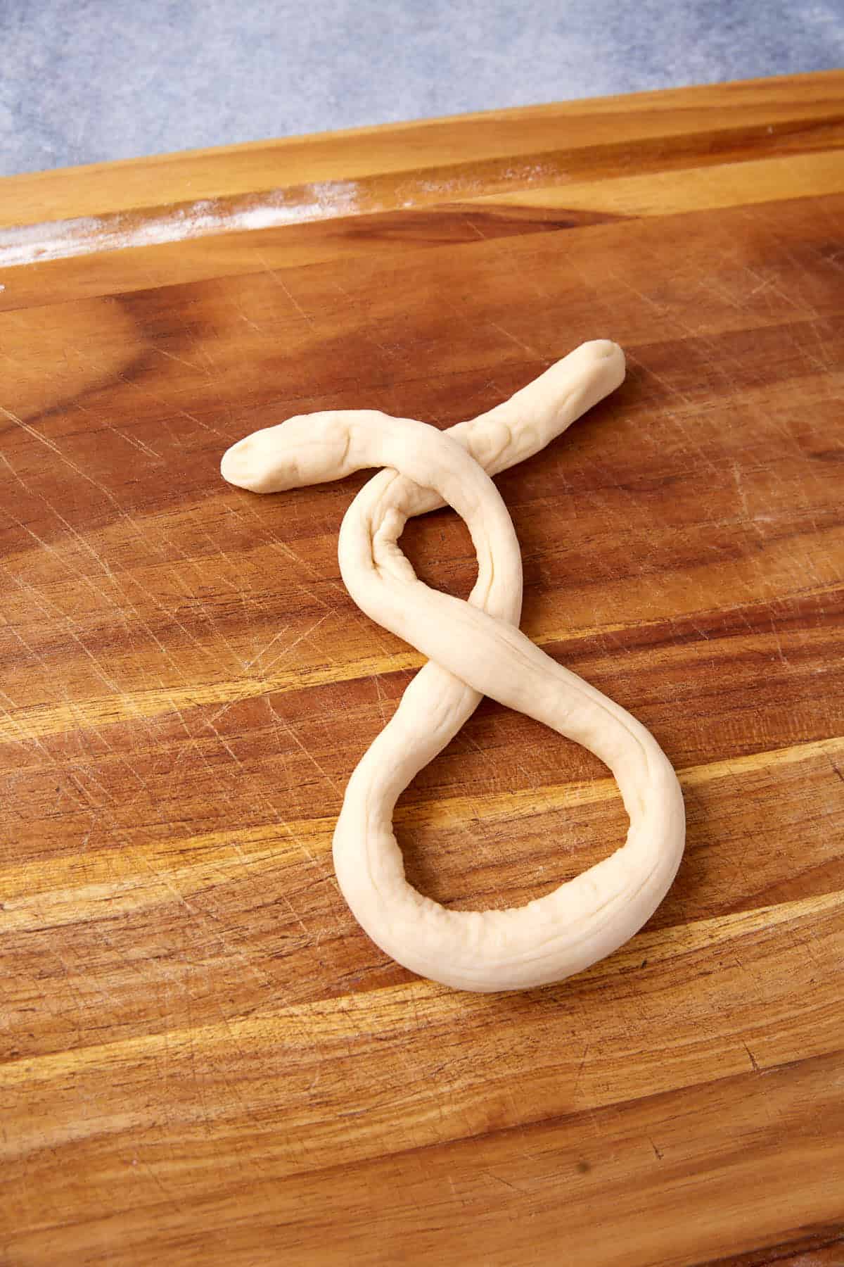A soft pretzel-shaped piece of dough forms a loose, twisted loop with ends pointing upward, resting on a wooden cutting board.