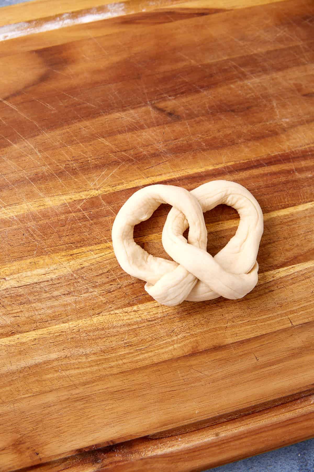 Unbaked soft pretzels in a twisted shape rest on a wooden cutting board with visible grain and scratch marks.