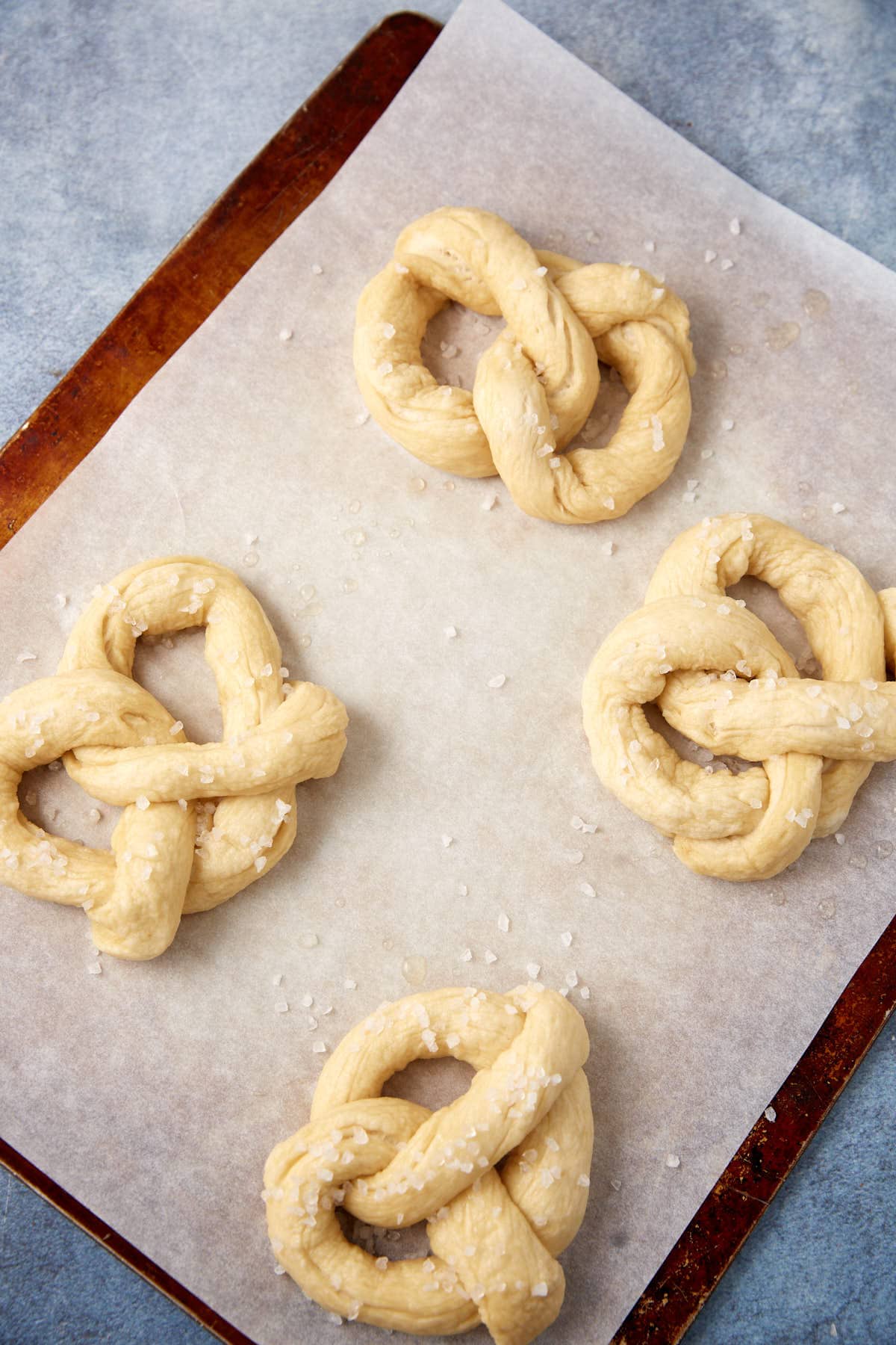 Four soft pretzels sprinkled with coarse salt are arranged on a parchment-lined baking sheet, ready to be baked. The background features a light blue surface.