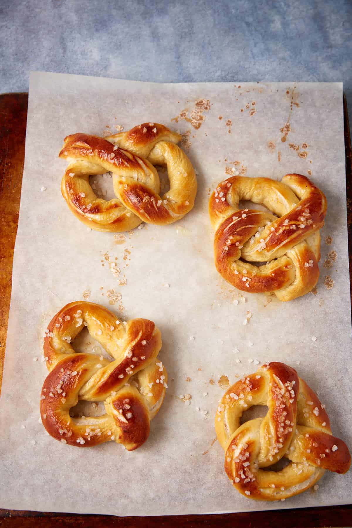 Four golden-brown soft pretzels sprinkled with coarse salt rest on parchment paper atop a wooden surface. The soft pretzels have a shiny, baked finish and are arranged in a loose, casual pattern.
