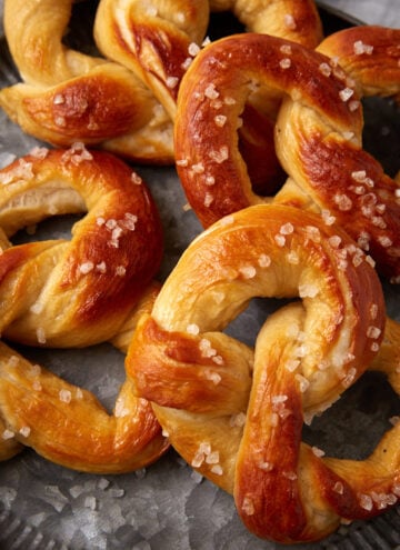 A close-up of soft pretzels with a golden-brown crust, topped with coarse salt, arranged on a gray metal plate.