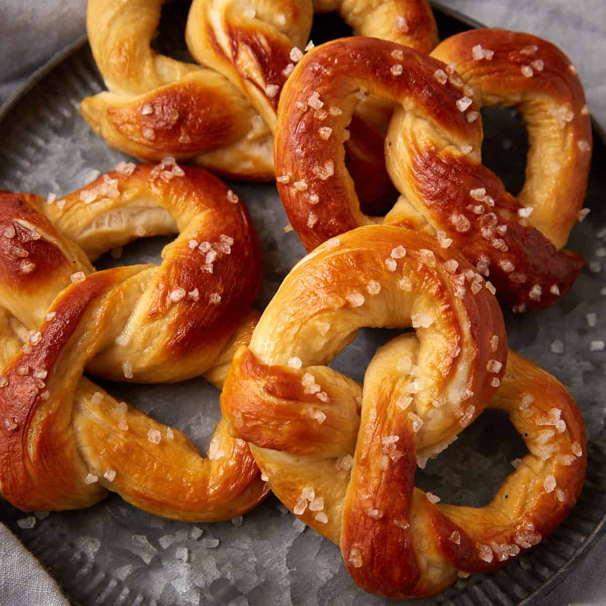 A close-up of soft pretzels with a golden-brown crust, topped with coarse salt, arranged on a gray metal plate.