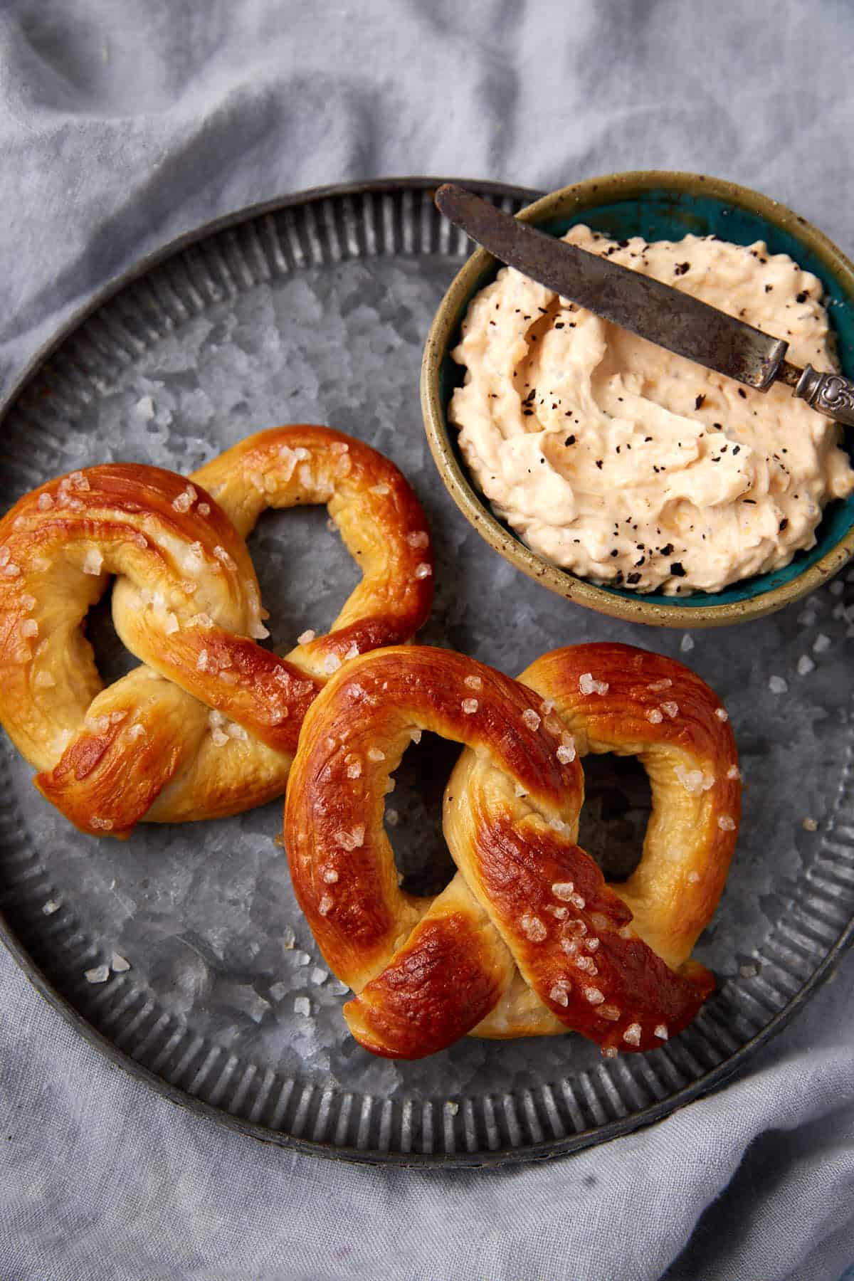 Two soft pretzels sprinkled with salt rest on a round metal tray beside a bowl of creamy dip topped with black seeds, a knife placed on the bowl. The tray sits atop a gray cloth.