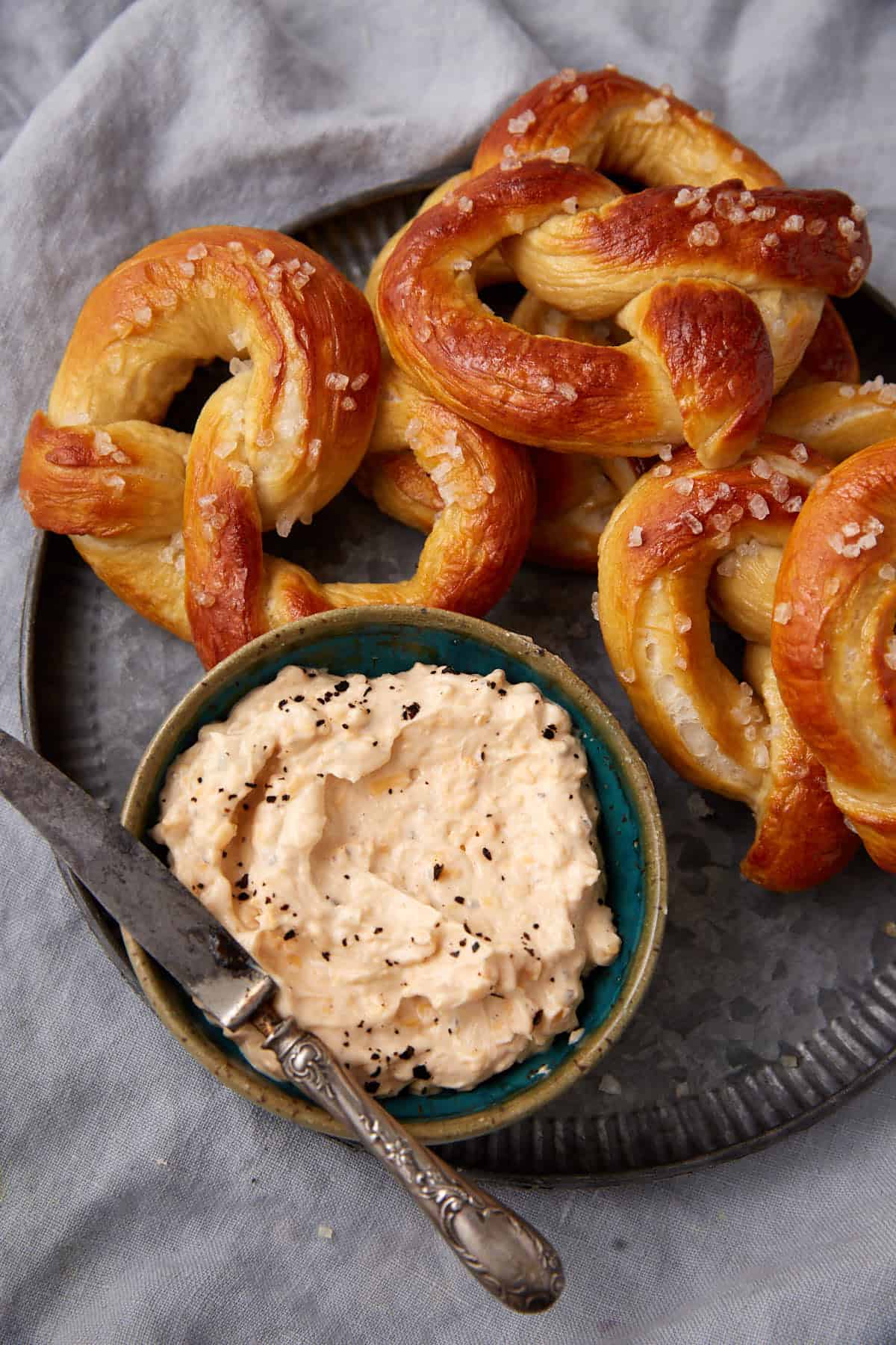 A plate with three soft pretzels and a bowl of creamy dip topped with black pepper; a butter knife rests on the side of the bowl, all set on a gray cloth.