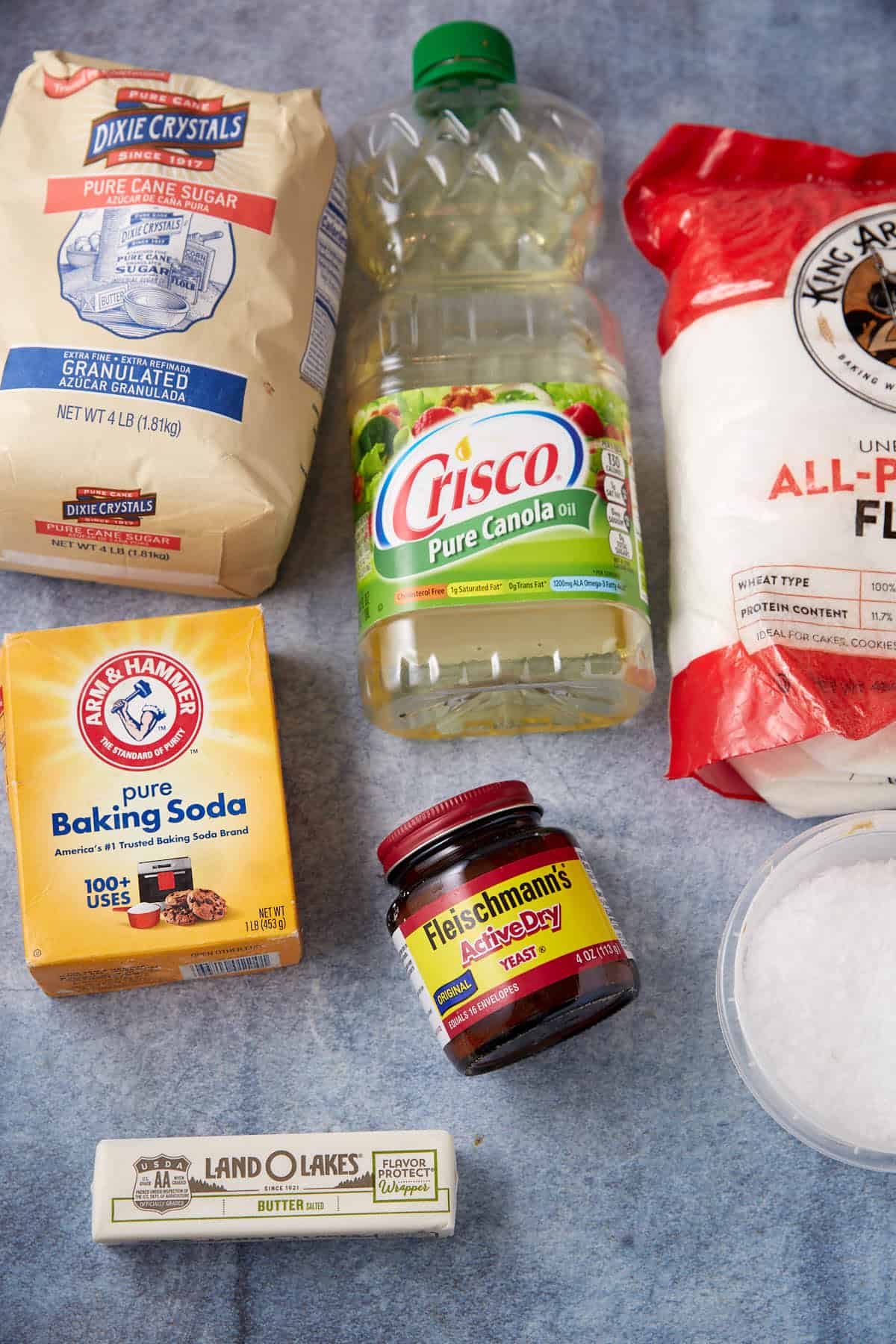 A collection of baking ingredients for soft pretzels, including granulated sugar, canola oil, all-purpose flour, baking soda, active dry yeast, a stick of butter, and a bowl of coarse salt on a blue surface.