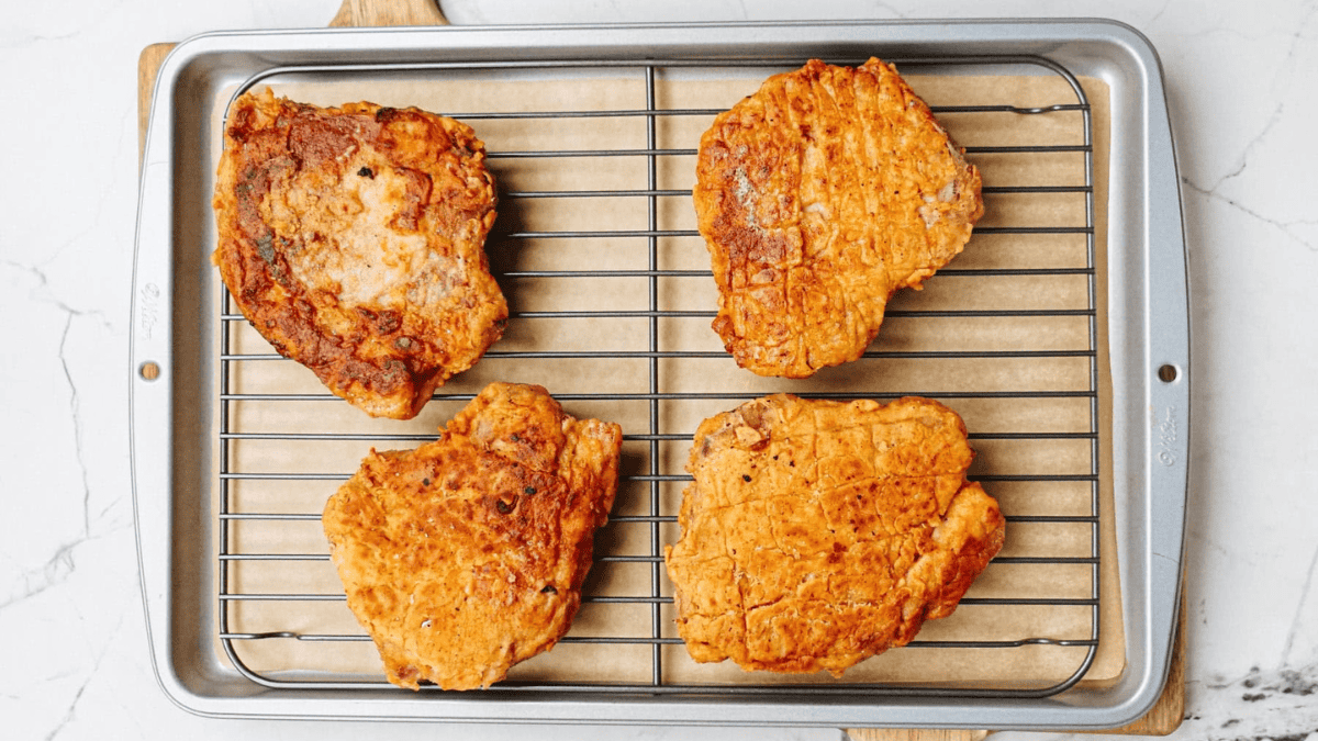 Four pieces of golden-brown fried chicken rest on a wire rack set over a baking sheet lined with parchment paper. The chicken pieces have a crispy, textured coating.