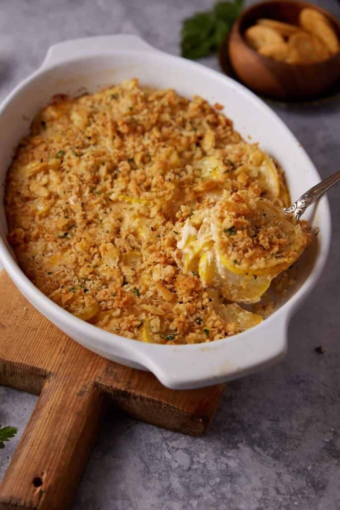 A white oval baking dish filled with yellow squash casserole, topped with a golden breadcrumb crust, sits on a wooden board. A serving spoon is lifting a portion out. In the background, there is a small bowl of crackers.