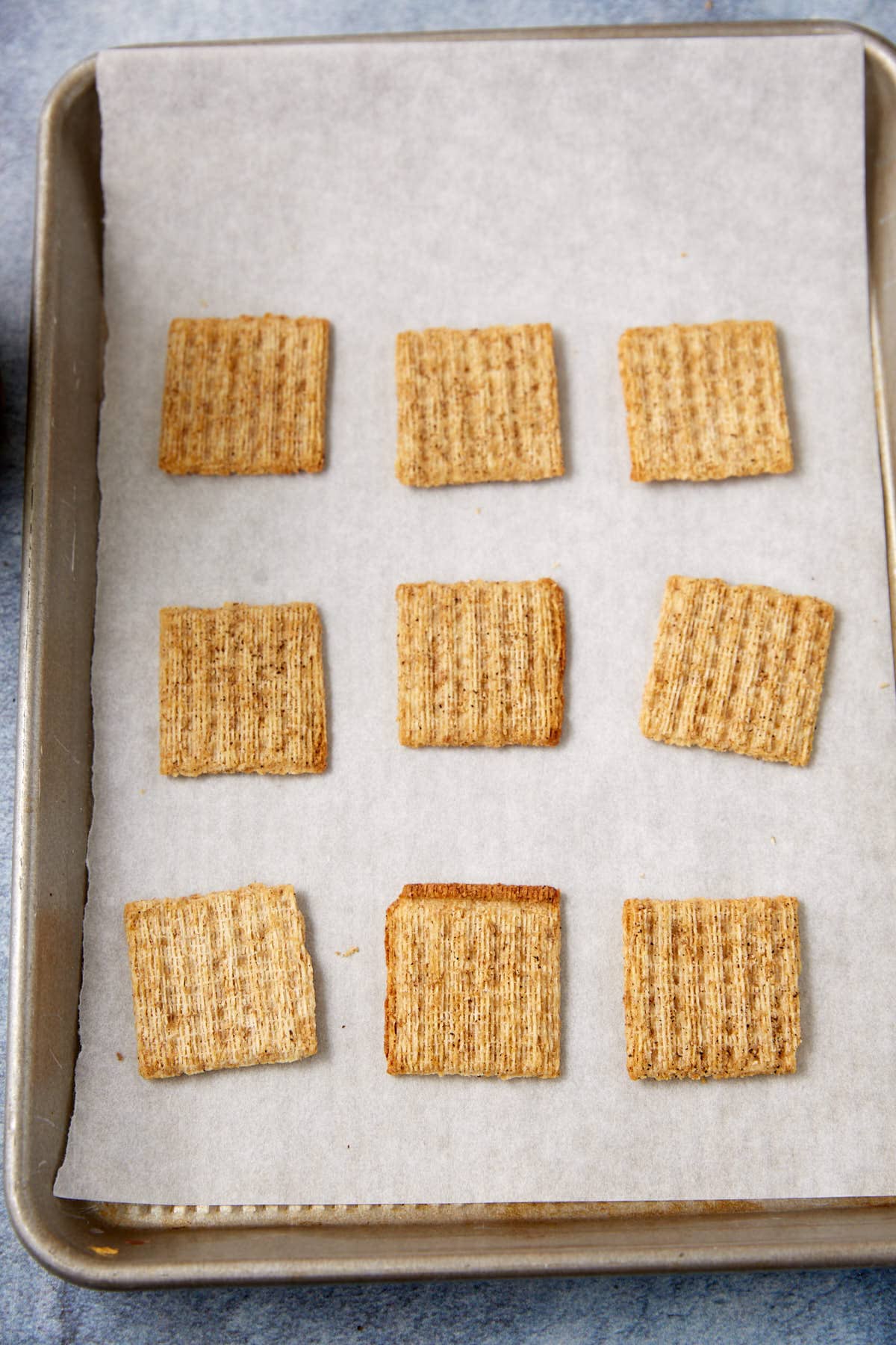 Nine square Dill Pickle Triscuit Appetizer crackers are arranged in three rows on a parchment-lined baking sheet with a light gray background. The crackers are evenly spaced and have a woven texture.