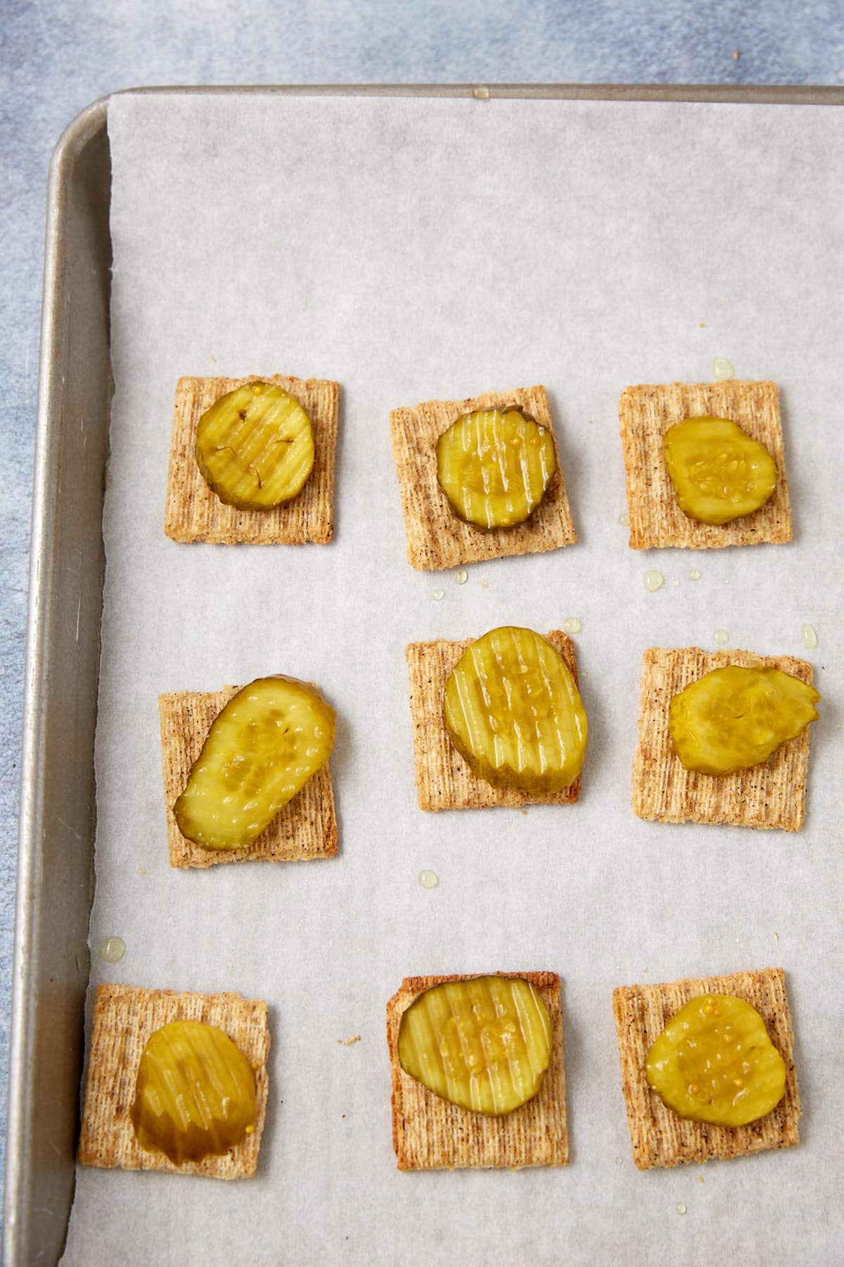 A baking tray lined with parchment paper holds nine Dill Pickle Triscuit Appetizer crackers, each topped with a round or oval pickle slice.