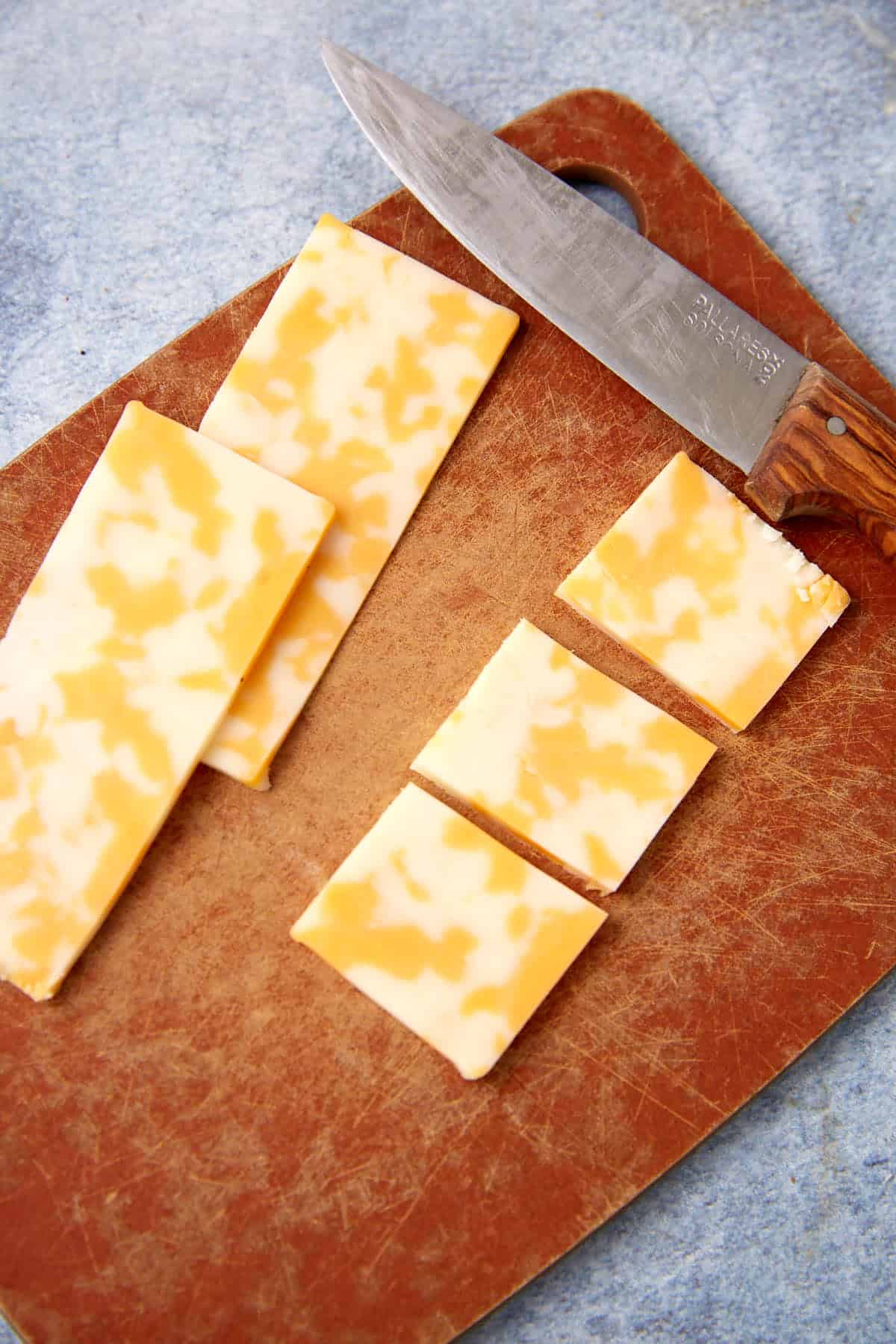 Sliced marbled Colby Jack cheese on a brown cutting board, with a wooden-handled knife resting nearby—perfect for assembling a Dill Pickle Triscuit Appetizer. The cheese is cut into both long strips and smaller square pieces.