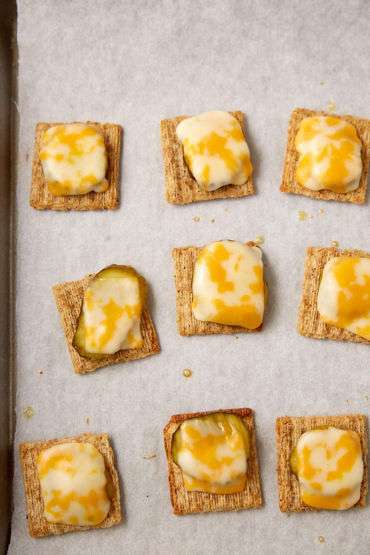 A baking sheet lined with parchment paper holds square wheat crackers topped with melted yellow and white cheese, and some feature pickle slices underneath, making it a tasty Dill Pickle Triscuit Appetizer.
