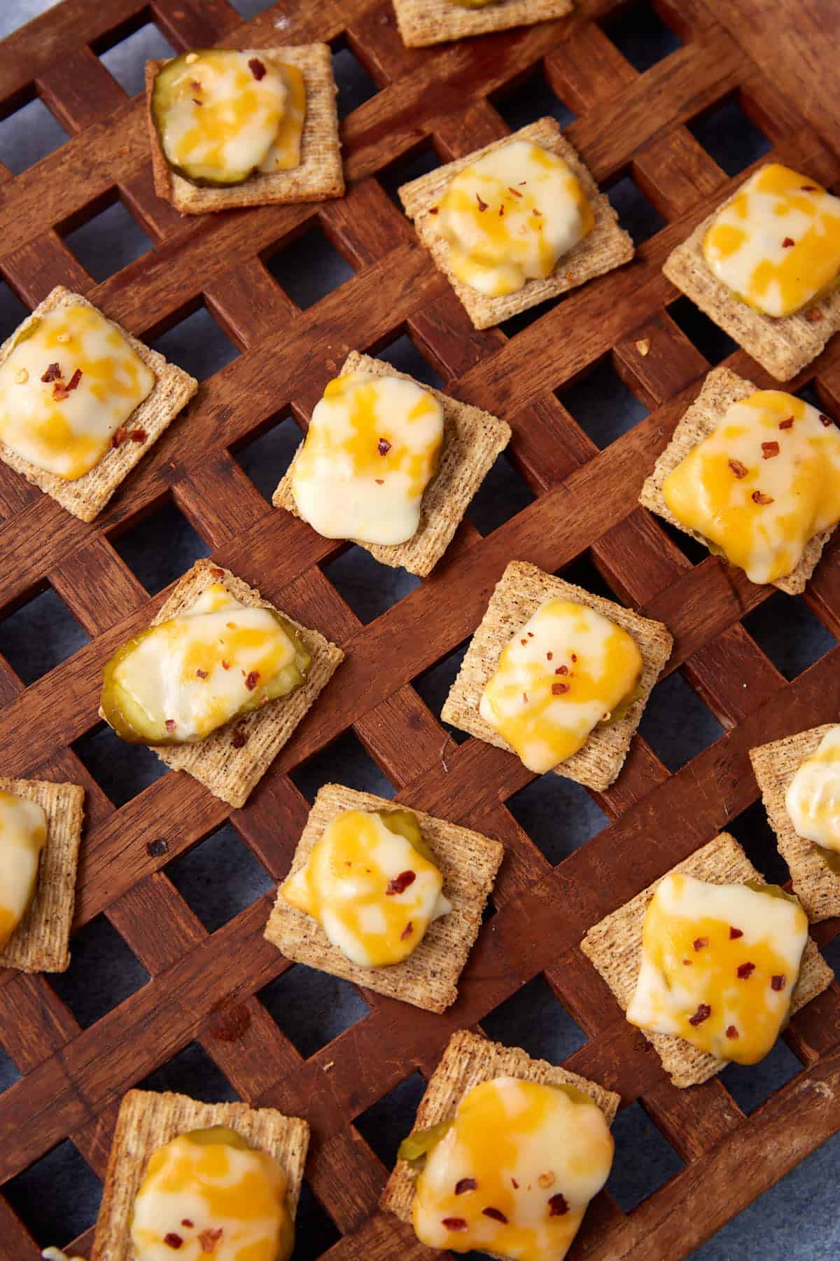 A lattice-style wooden tray holds a Dill Pickle Triscuit Appetizer&mdash;square wheat crackers topped with melted yellow and white cheese; some are finished with pickle slices and red pepper flakes.