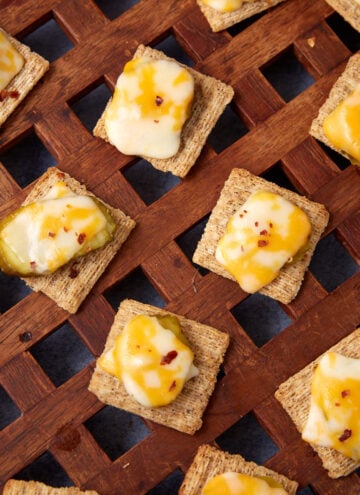 Several woven wheat crackers, in this Dill Pickle Triscuit Appetizer, are topped with melted cheese, sliced pickles, and red pepper flakes, artfully arranged on a wooden lattice surface.