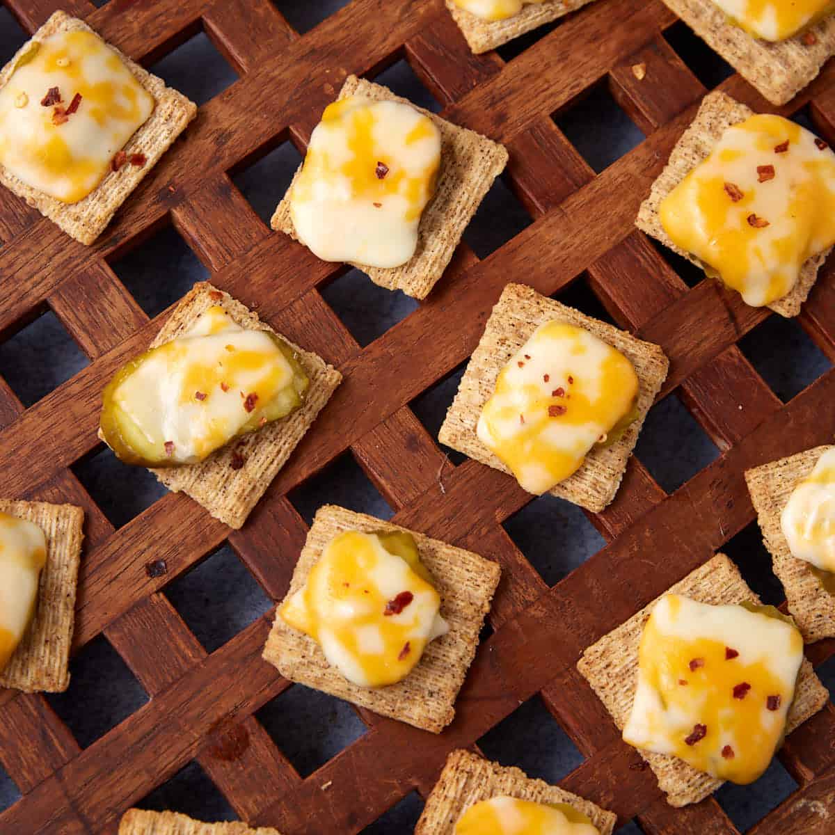 Several woven wheat crackers, in this Dill Pickle Triscuit Appetizer, are topped with melted cheese, sliced pickles, and red pepper flakes, artfully arranged on a wooden lattice surface.