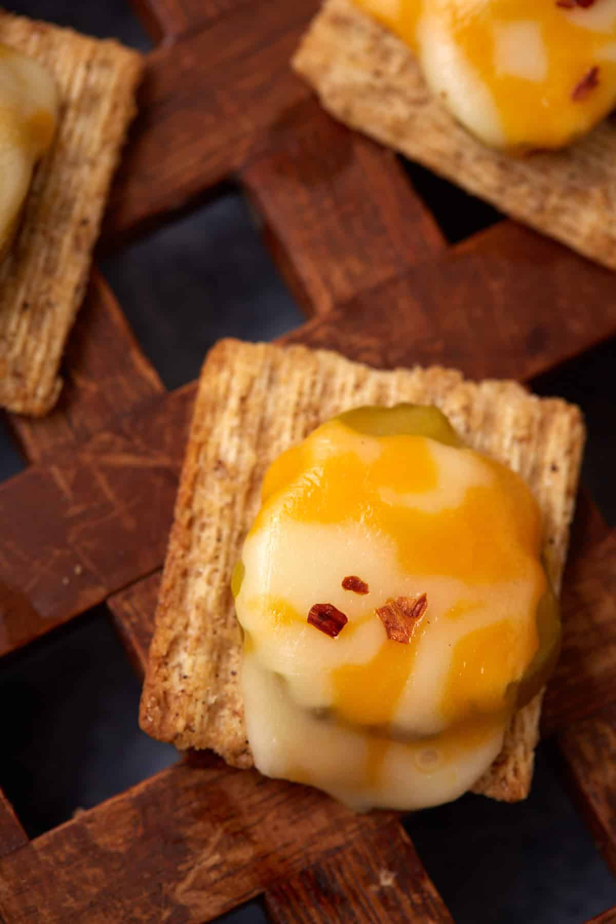A close-up of a Dill Pickle Triscuit Appetizer topped with melted cheddar and mozzarella cheese, a slice of jalapeño, and a sprinkle of red pepper flakes on a wooden surface.