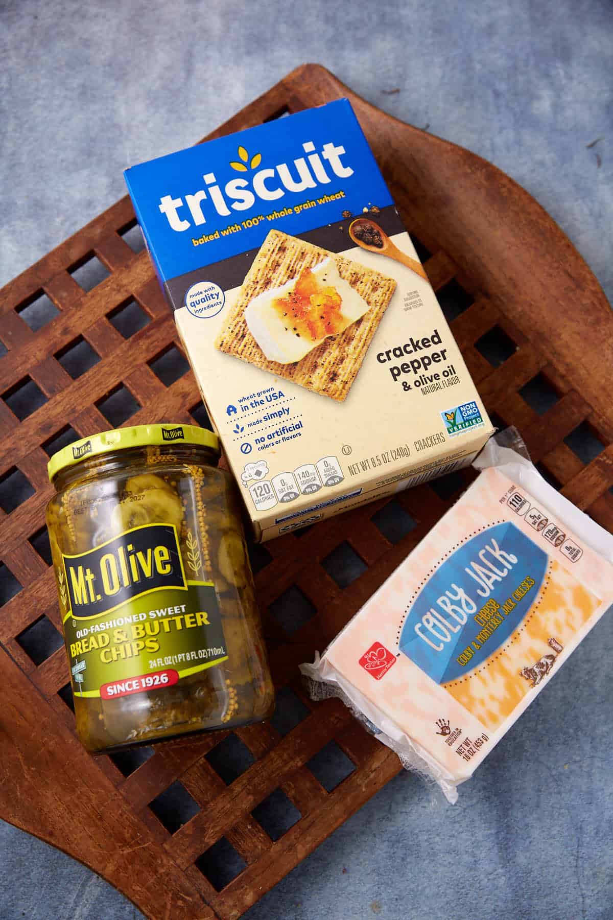 A tasty Dill Pickle Triscuit Appetizer: Mt. Olive bread and butter chips, Triscuit cracked pepper & olive oil crackers, and Colby Jack cheese served on a wooden tray with a blue background.