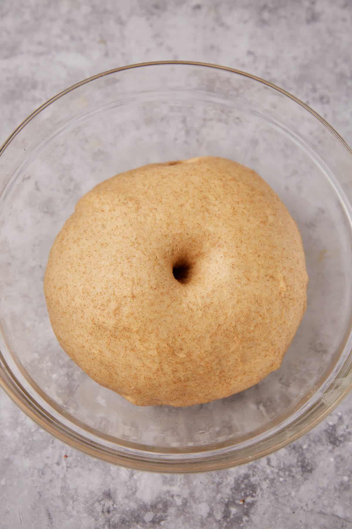 A ball of dough for whole wheat rolls with a poke mark in the center sits in a clear glass bowl on a gray, textured surface.