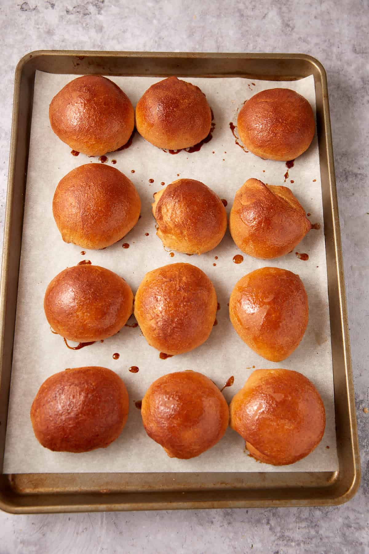 A baking tray lined with parchment paper holds twelve golden brown, freshly baked whole wheat rolls arranged in neat rows on a light gray surface.
