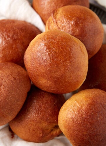 A close-up of several golden-brown, shiny whole wheat rolls piled together on a white cloth.