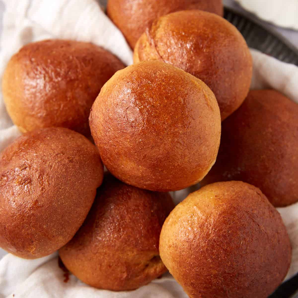 A close-up of several golden-brown, shiny whole wheat rolls piled together on a white cloth.