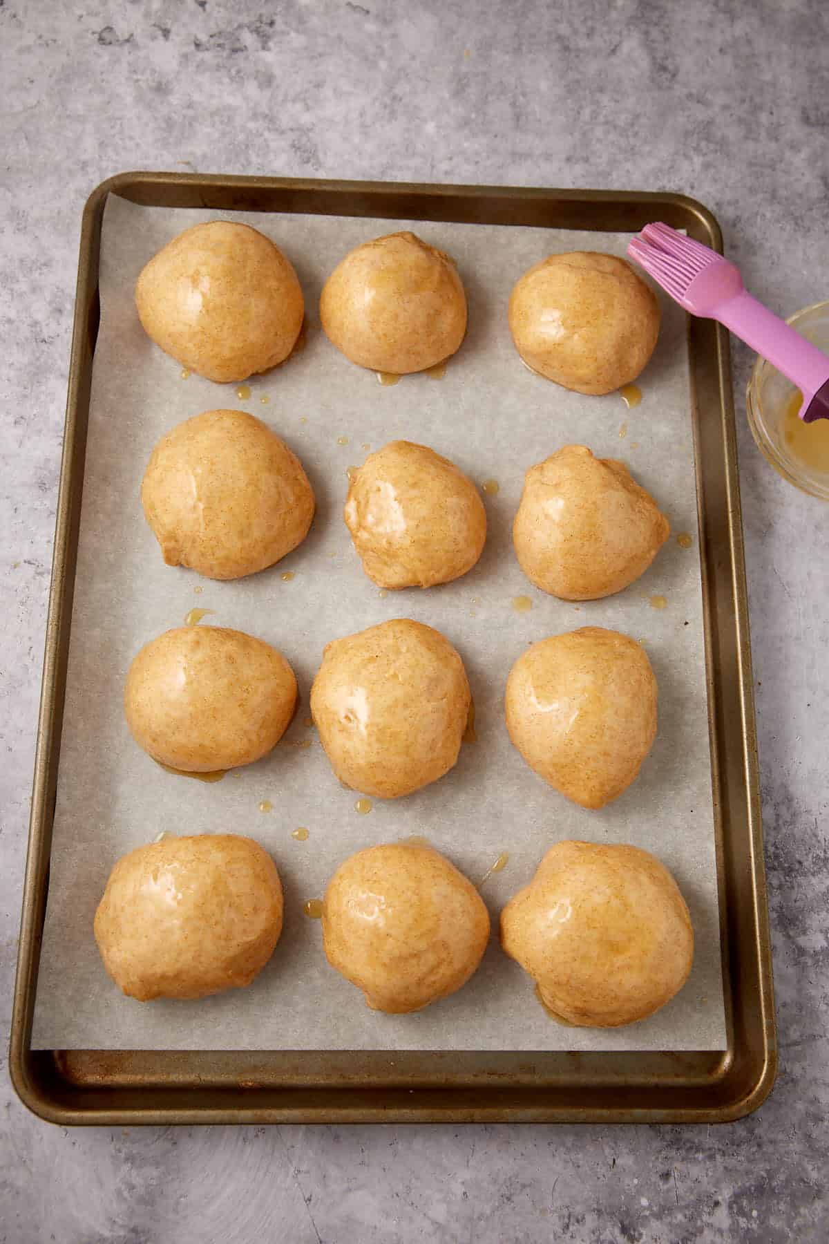 Twelve round whole wheat rolls brushed with egg wash are arranged on a parchment-lined baking sheet, with a pink pastry brush and a small bowl next to them on a gray surface.