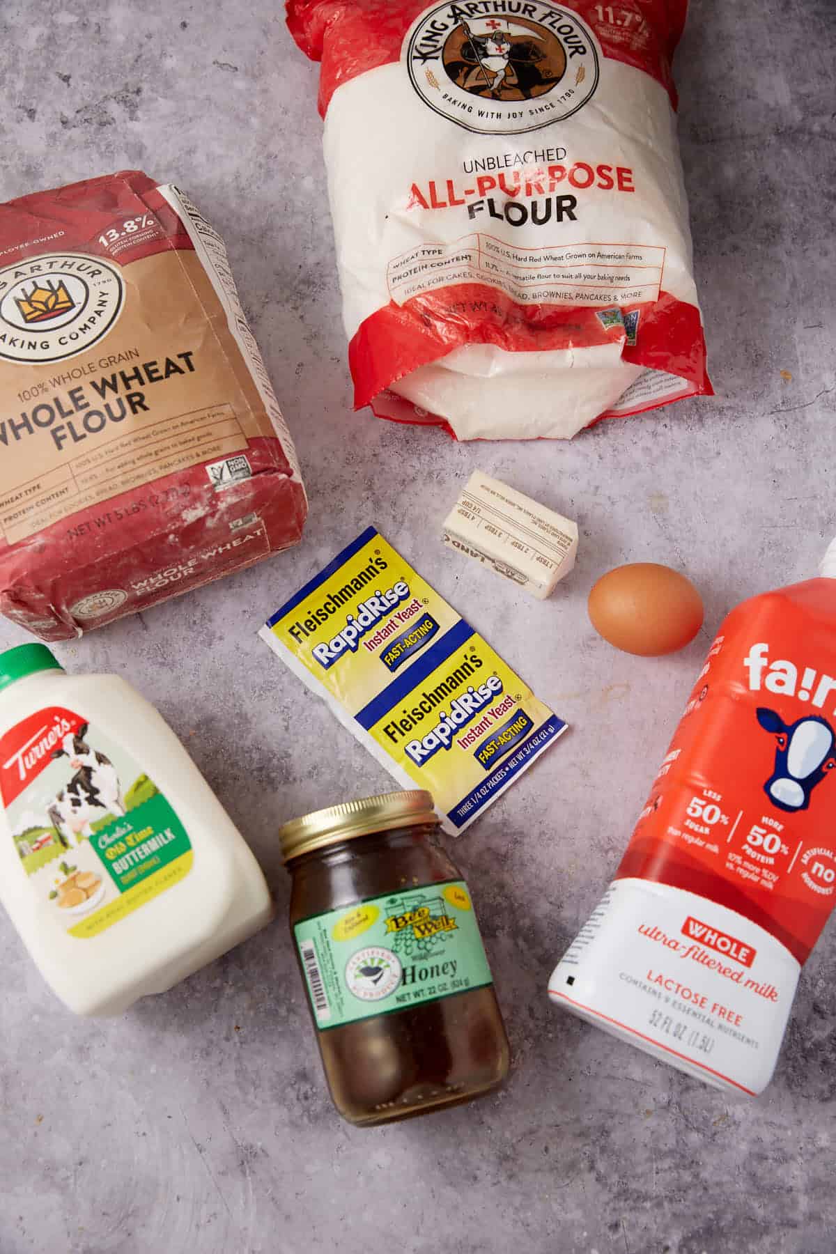 Assorted baking ingredients on a gray surface: bags of whole wheat and all-purpose flour, sticks of butter, yeast packets, a brown egg, a milk jug, jar of honey, and lactose-free milk—everything you need to bake delicious whole wheat rolls.