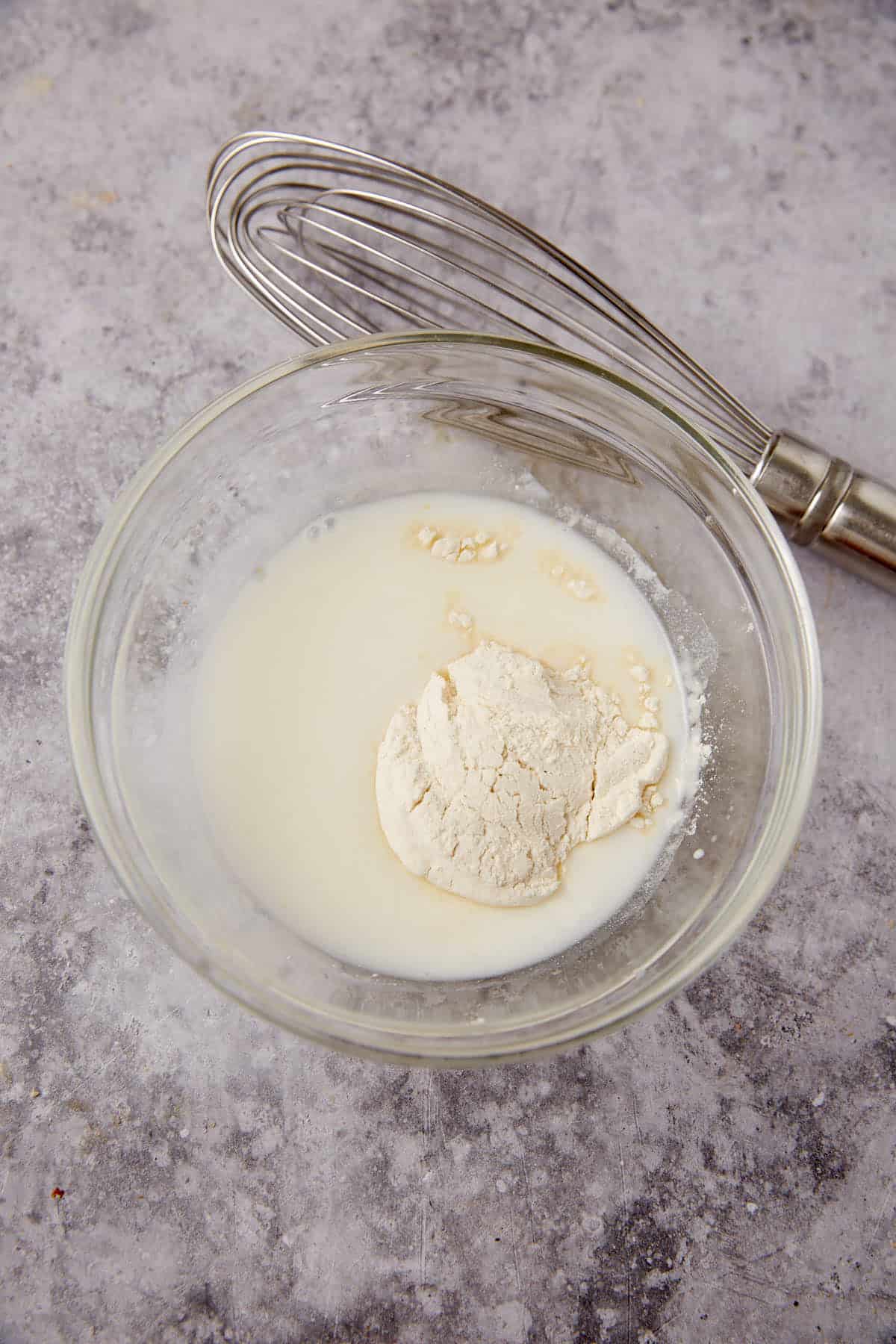A glass bowl containing milk and flour, perfect for making whole wheat rolls, sits on a gray countertop with a metal whisk placed nearby.