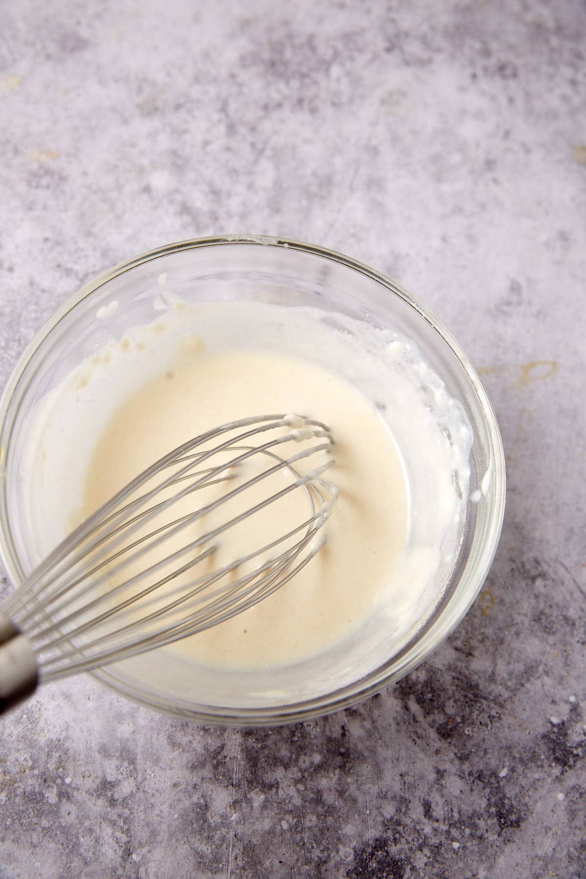 A glass bowl filled with creamy white batter is being whisked with a metal whisk on a textured, light gray surface, perfect for making soft whole wheat rolls.
