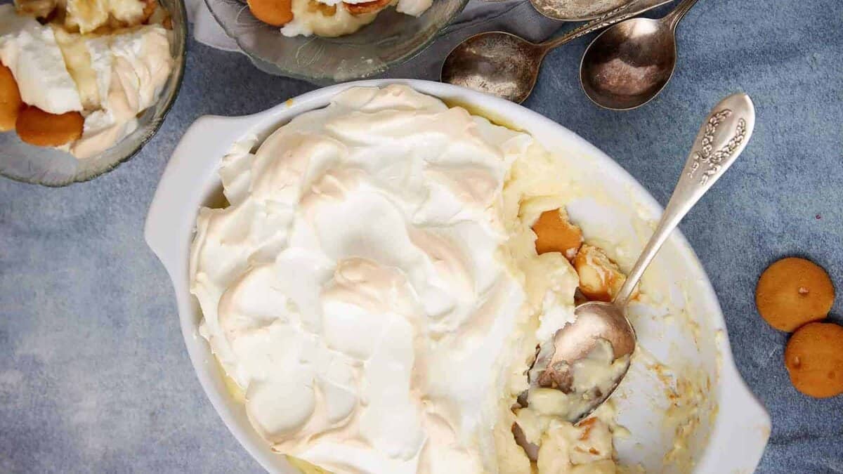 A dish of banana pudding topped with meringue, partially served with a spoon inside, surrounded by small bowls of pudding and vintage spoons, just like I followed Grandma’s 1950s recipe cards on a blue surface.
